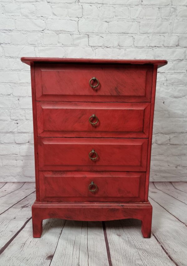 A small red wooden dresser with four drawers and brass ring handles stands on a wooden floor against a white brick wall background.