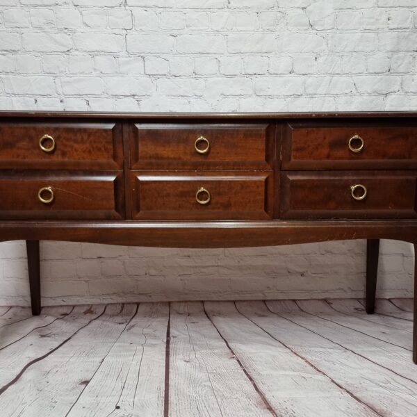 A dark wood sideboard with six drawers, each featuring a gold circular handle, stands against a white brick wall. The sideboard has long, slender legs and a smooth finish, with the wood's grain and texture visible. The floor is made of distressed wood planks.