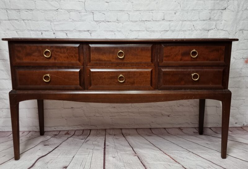 A dark wood sideboard with six drawers, each featuring a gold circular handle, stands against a white brick wall. The sideboard has long, slender legs and a smooth finish, with the wood's grain and texture visible. The floor is made of distressed wood planks.