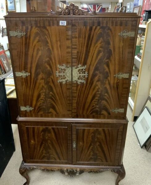 A tall, antique wooden cabinet with ornate metal accents and detailed carving at the base is displayed in a room filled with various other pieces of furniture and framed artwork. The cabinet features two large doors on the upper section and two smaller doors on the lower section.