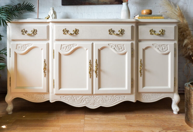 A cream-colored, vintage sideboard with ornate gold handles and intricate carvings sits on a hardwood floor. The sideboard features three drawers and four cabinet doors. It is flanked by lush plants and decorated with books, a candle, and a small sculpture on top.