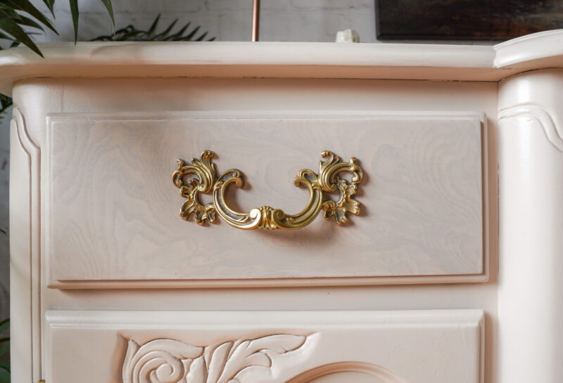 A close-up of a vintage, light-colored dresser drawer featuring an ornate gold handle. The drawer front has intricate carved details, and there are some plants in the blurred background.