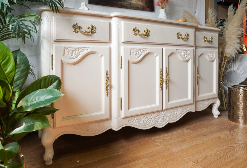 An ornate, antique-style white sideboard with intricate gold handles and decorative carvings on the doors and drawers. It stands on wooden flooring beside various green plants and eclectic home decor items.