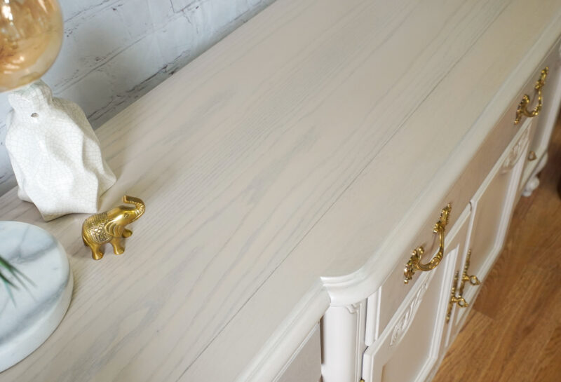 A top view of a light-colored wood dresser with brass handles. On the dresser are decorative items, including a white textured vase, a small golden elephant figurine, and a glass orb on a marble base. The backdrop is a light gray brick wall.