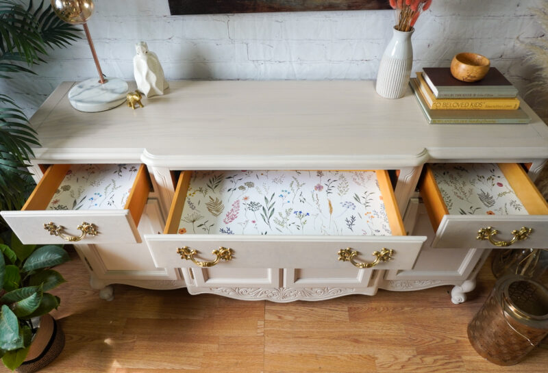 A light-colored wooden dresser with six floral-patterned lined drawers. Three top drawers are opened, revealing the lining. Decorations, including a statue, books, a vase, and a bowl, are on top. It's surrounded by green plants and has a wooden floor base.