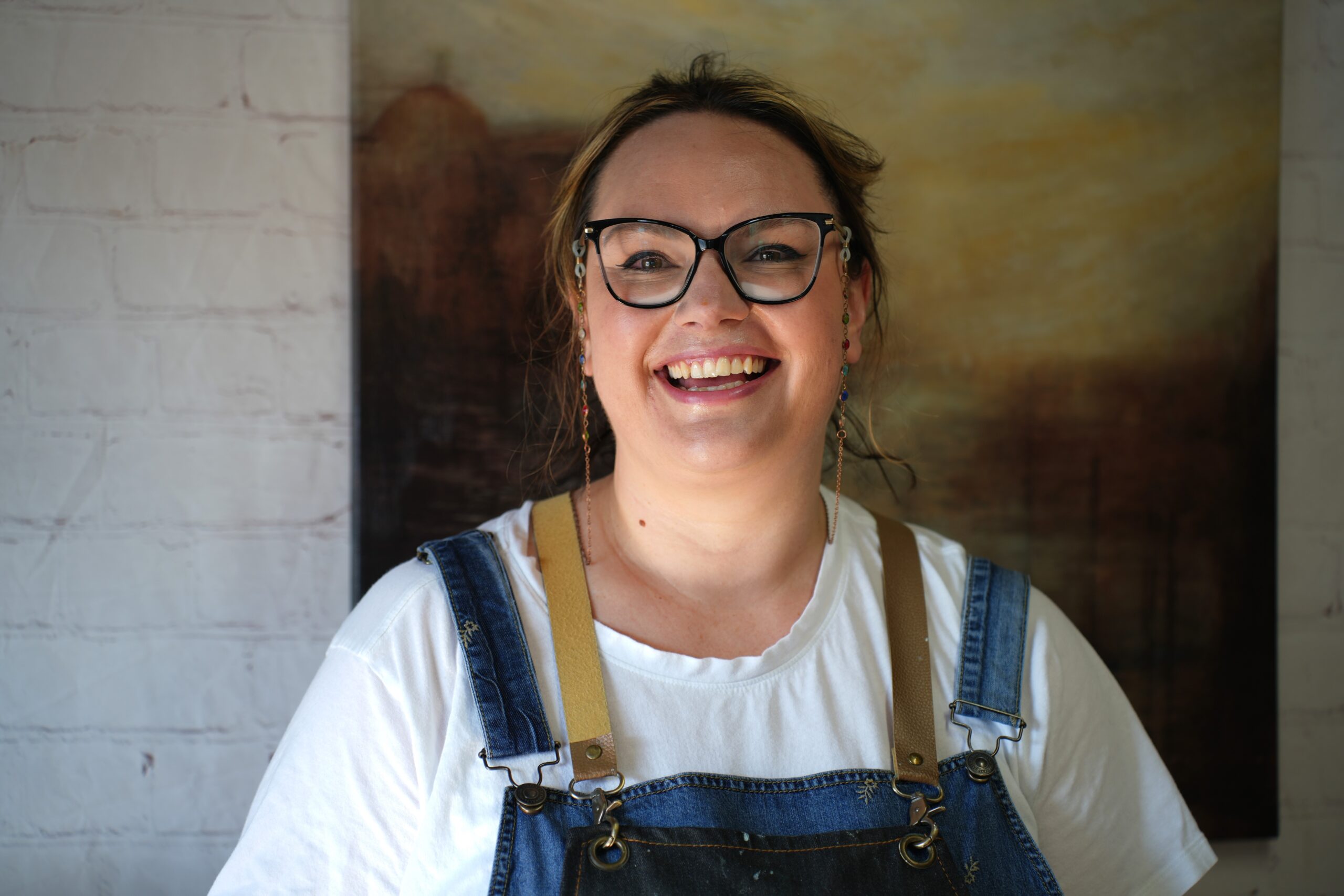 A person with light brown hair in a ponytail is smiling widely. They are wearing glasses, a white shirt, and blue denim overalls. The background shows a brick wall and a framed painting with earthy tones.