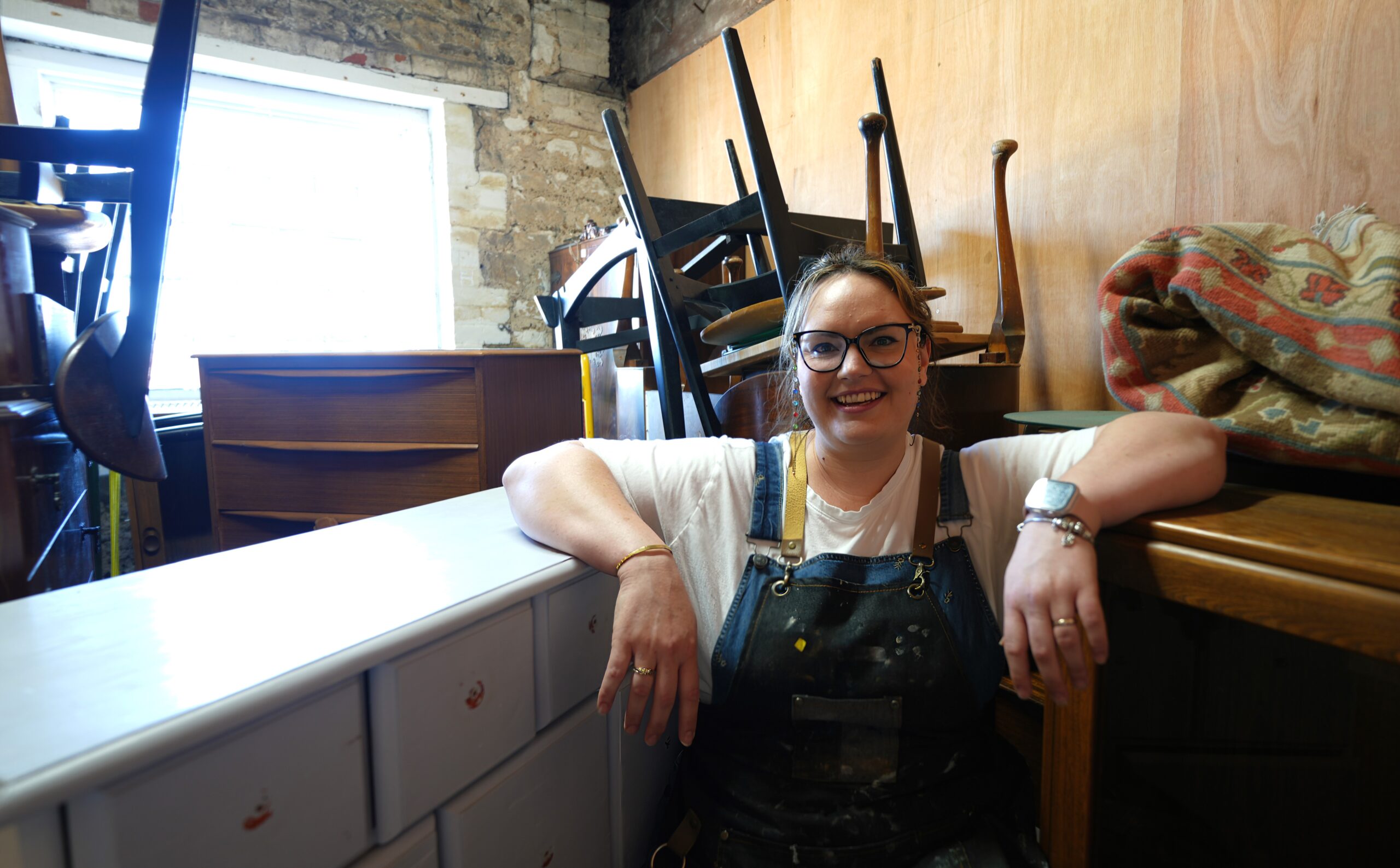 A woman wearing glasses and a white shirt under overalls leans on a white dresser in a workshop filled with stacked chairs and various pieces of furniture. A window on the left side emits natural light, illuminating the space. She smiles at the camera.