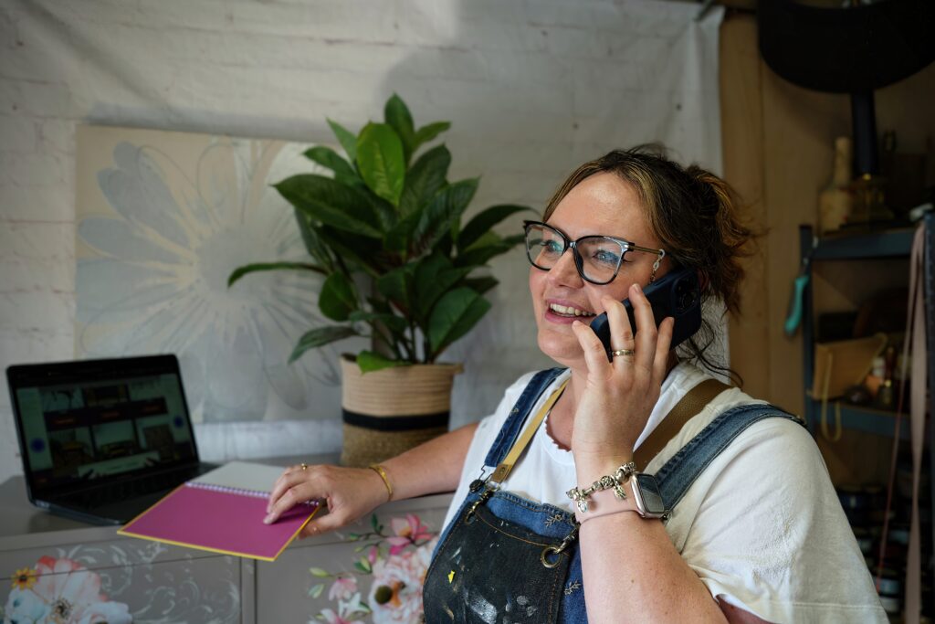 A woman wearing glasses and overalls, smiling while talking on a phone. She stands at a desk with one hand on a pink notebook and a laptop open in the background. A potted plant and various items can be seen in the room.