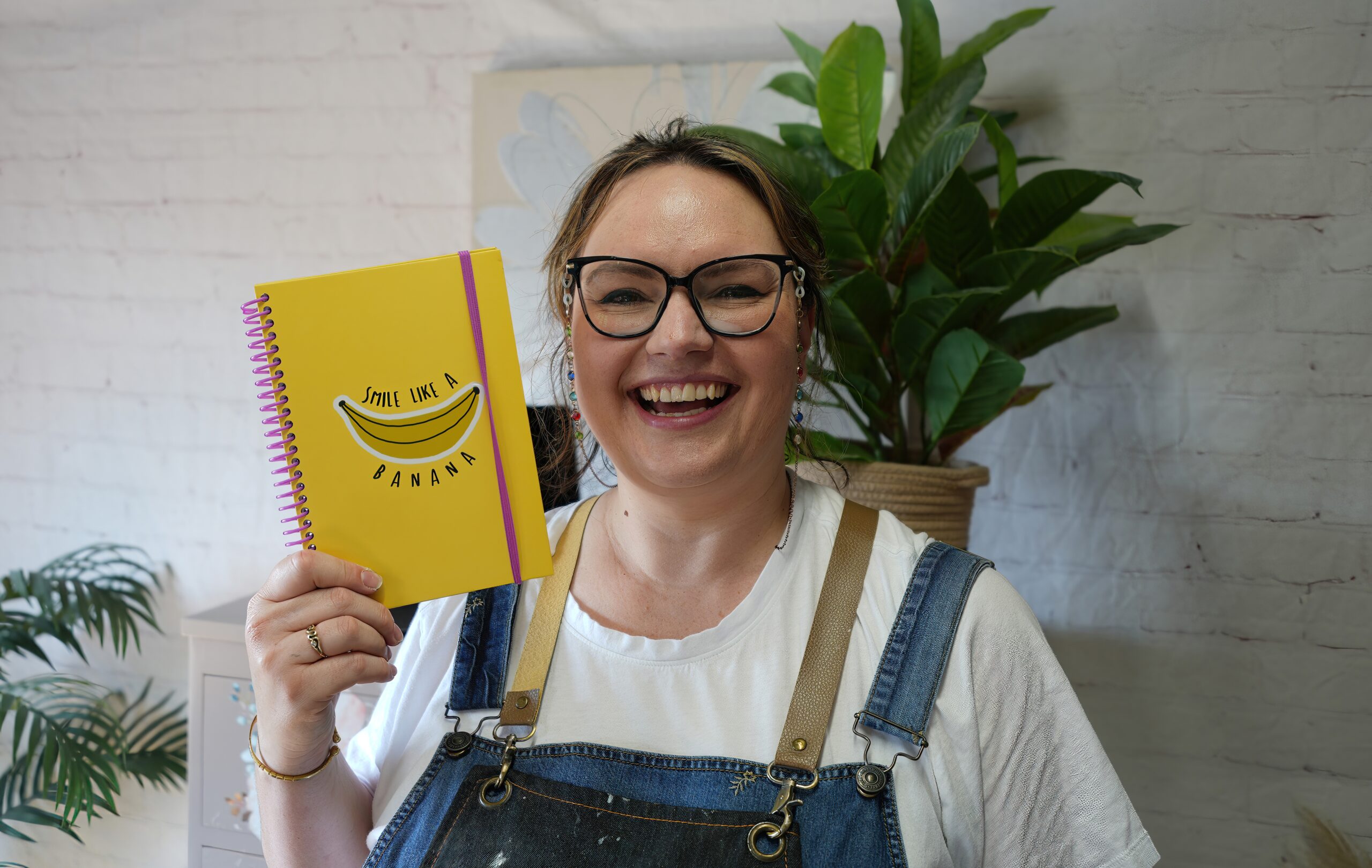 A person with glasses and a big smile stands indoors holding a yellow notebook with the words "Smile Like A Banana" on the cover. They are wearing a white shirt under denim overalls. There is a plant in the background.