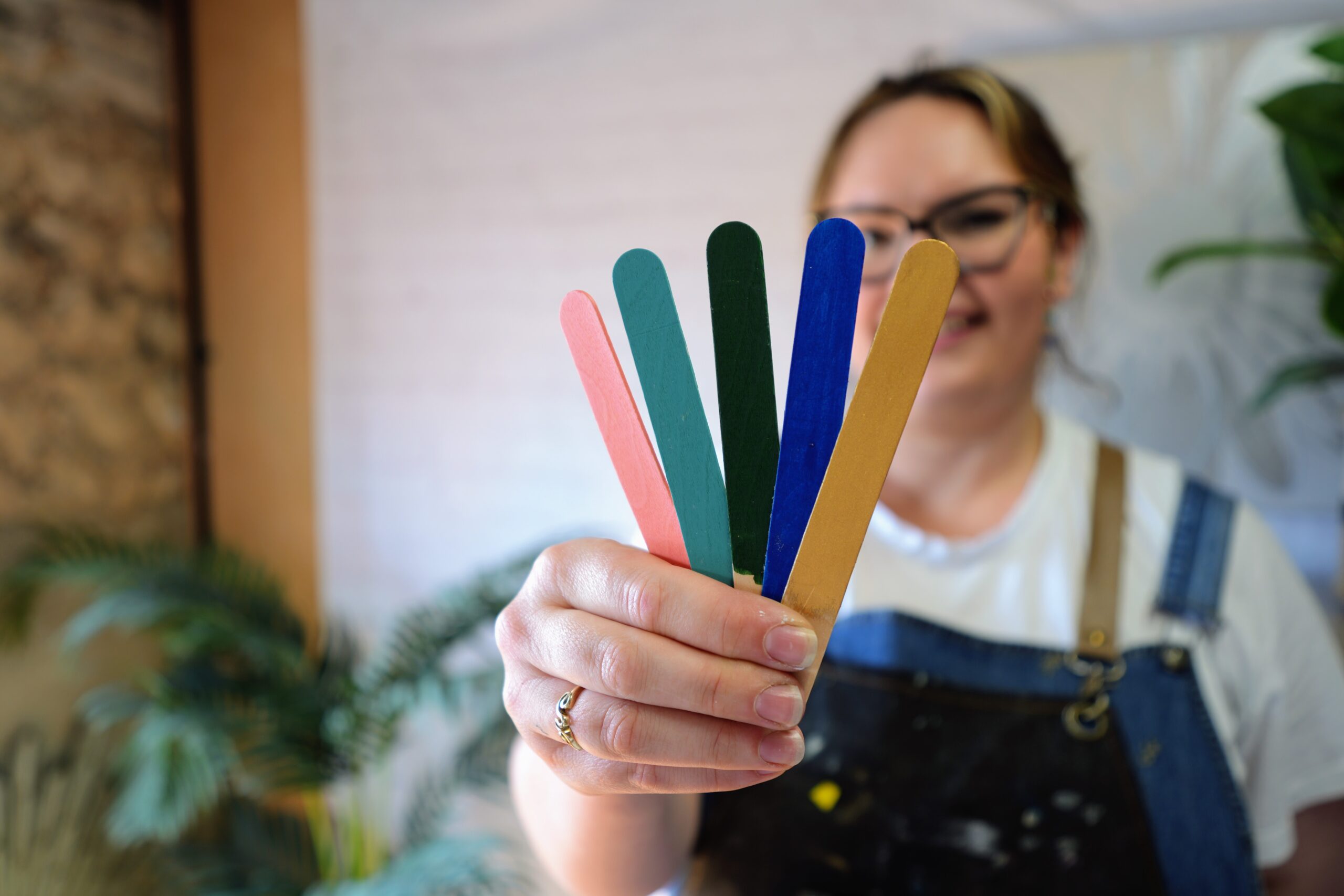 A person in glasses and overalls holds up five colorful wooden sticks, displaying them in front of the camera. The sticks are red, green, blue, and two different shades of yellow. The background includes indoor plants and neutral-colored walls.