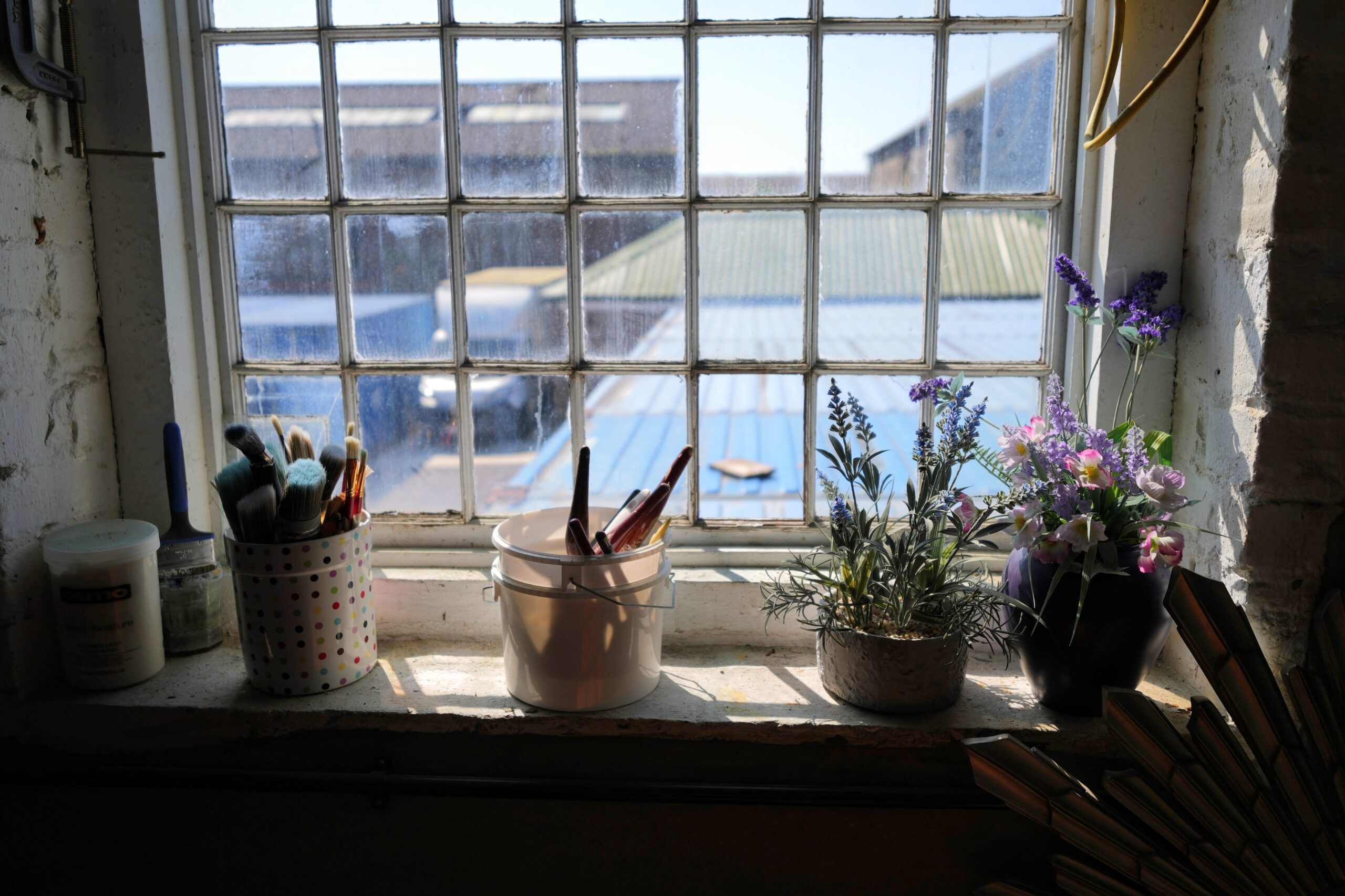 An artist's window sill featuring jars of paintbrushes and containers of artificial flowers, illuminated by sunlight streaming through a multi-pane window. The scene outside includes industrial buildings with blue and green rooftops.