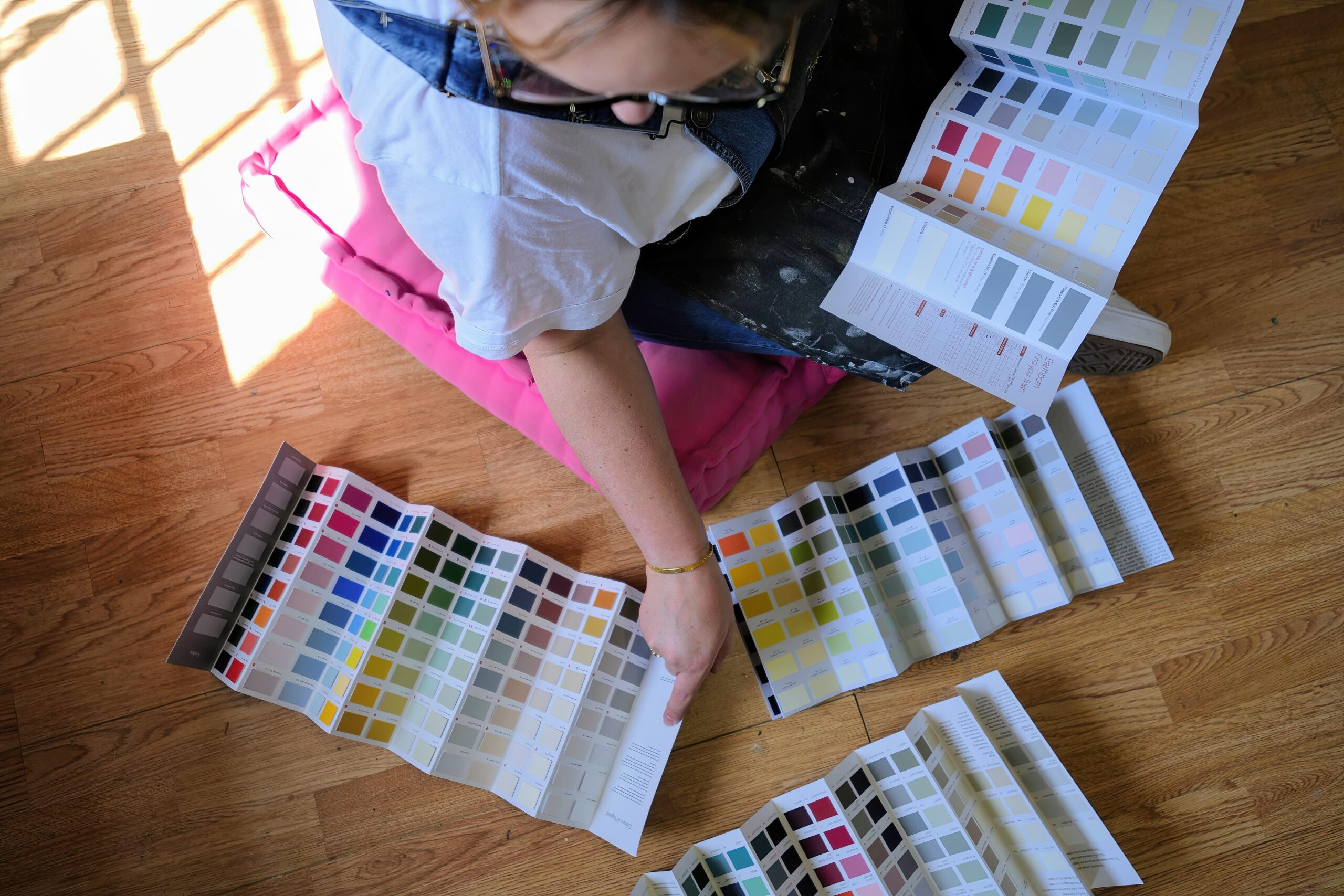 A person wearing glasses and overalls sits on a pink pillow on a wooden floor, looking at and pointing to large color swatches. The scene is bathed in natural light from a window.