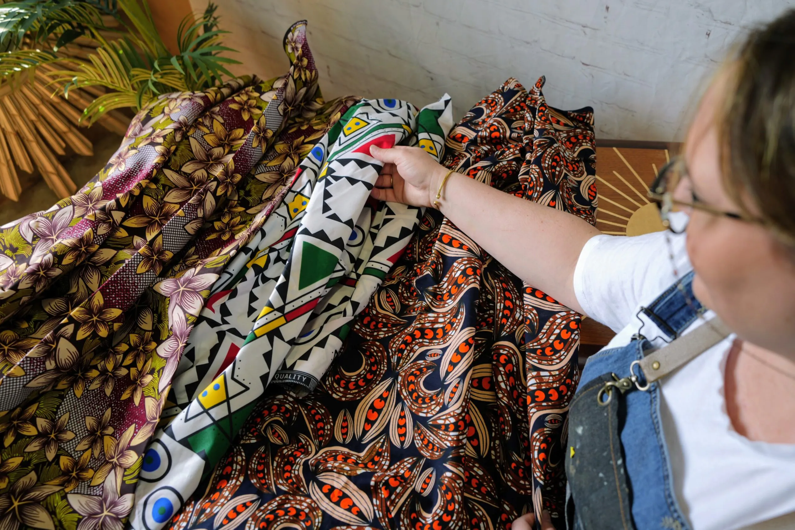 A person wearing glasses and an apron is examining various colorful fabric swatches with vibrant patterns, including floral and geometric designs. The fabrics are spread out on a wooden surface, with some greenery in the background.