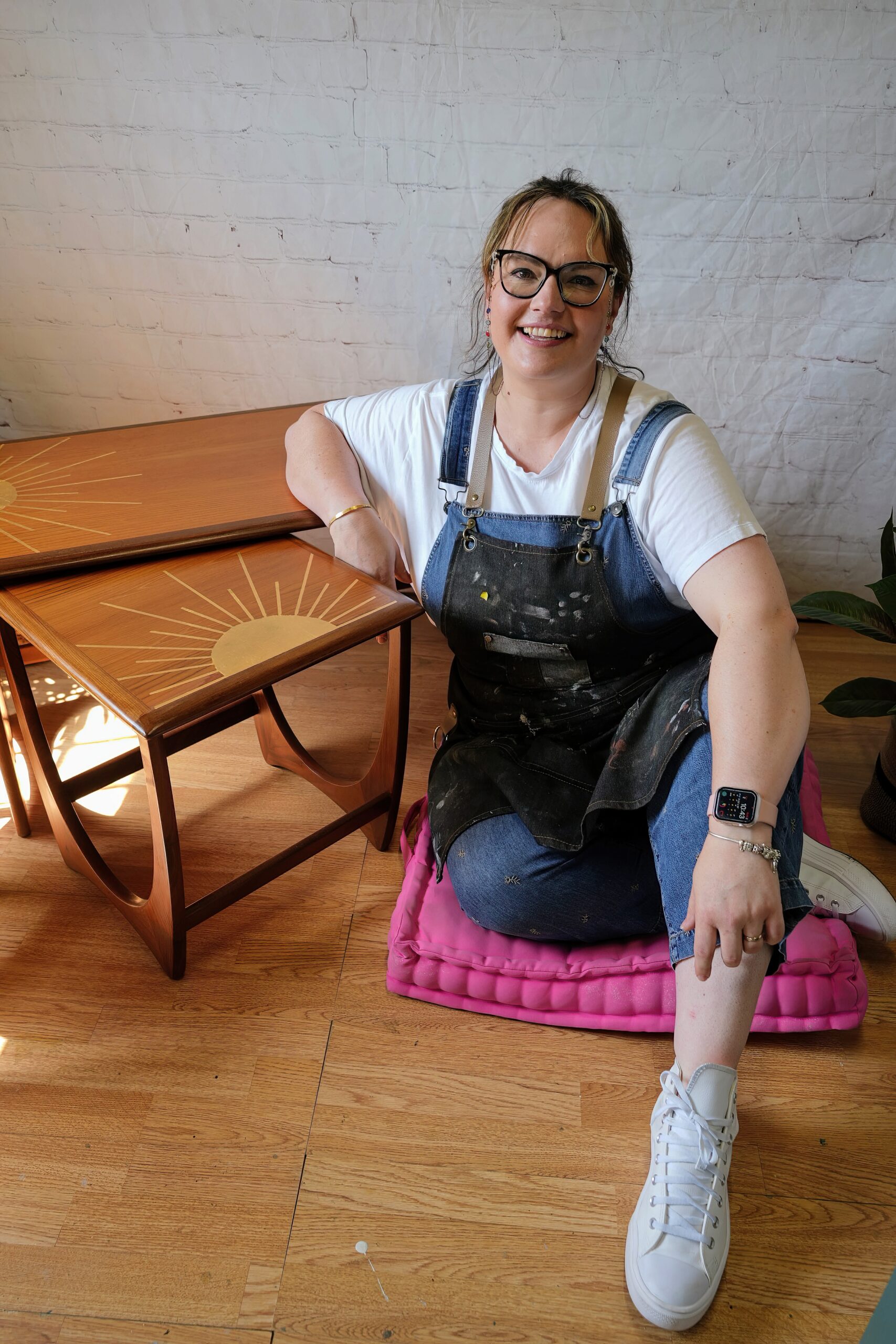 A person with glasses and light hair, wearing a white shirt, overalls, and a black apron, sits on a pink cushion on a wooden floor. They are smiling and resting their arm on a wooden table with a sunburst design. A potted plant is partially visible on the right.