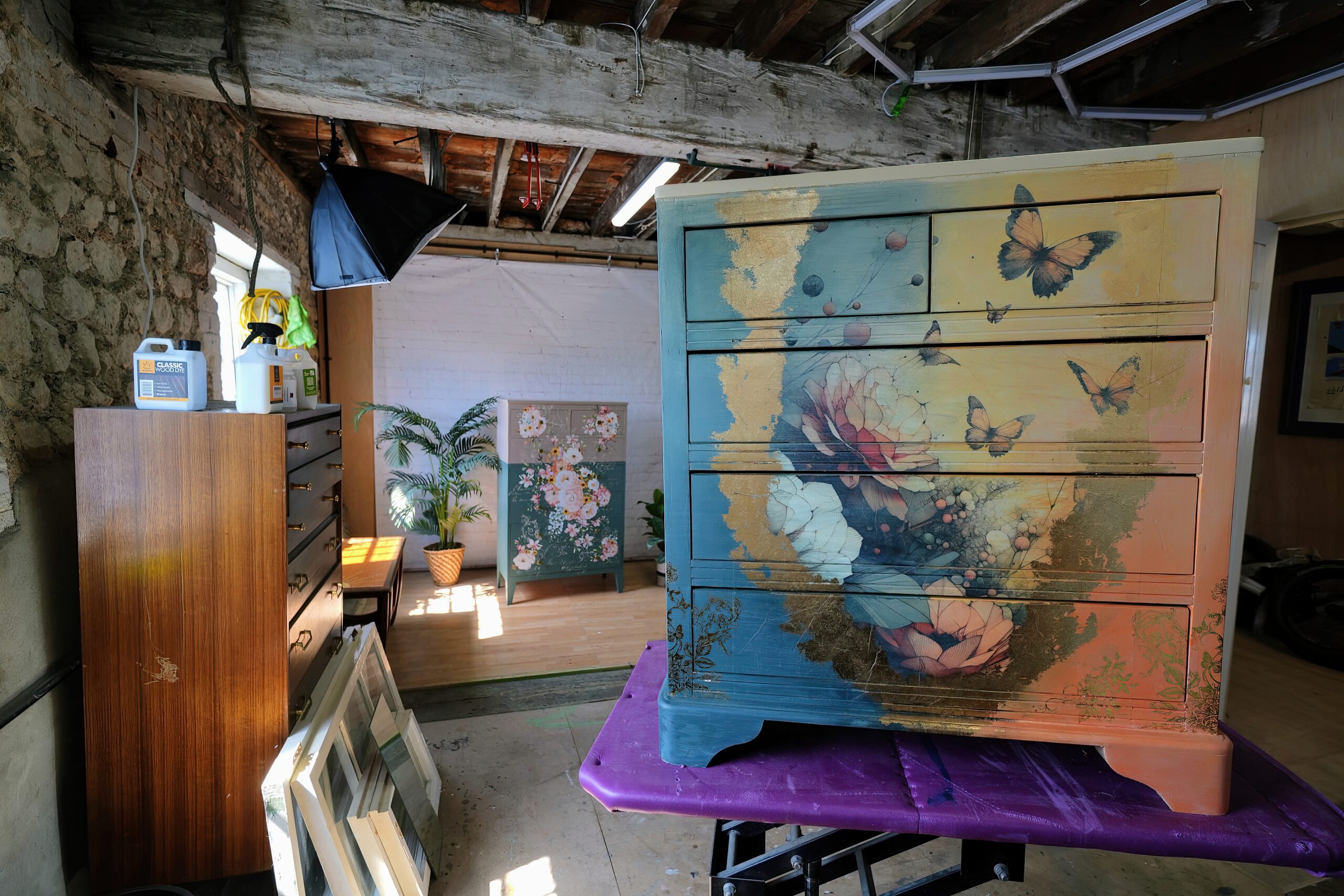 A room with rustic wooden beams and stone walls houses a painted wooden dresser adorned with floral and butterfly motifs at the forefront on a purple cloth. In the background, another floral painted dresser and a potted palm plant are visible.