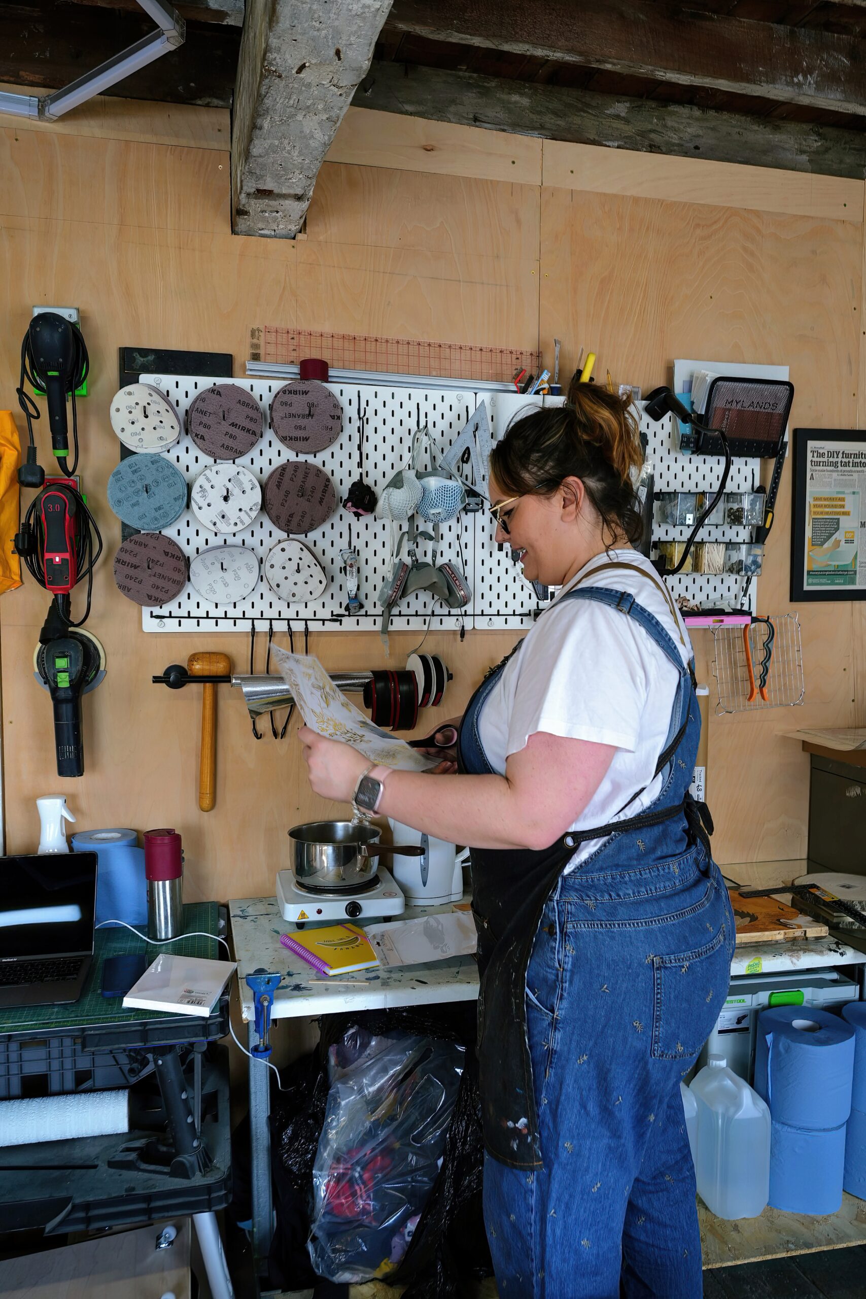 A person wearing denim overalls and a white t-shirt reads a paper in a workshop. The workshop has tools, sandpaper, and supplies hanging on a pegboard, along with various other items on a worktable. The person appears focused on their task.