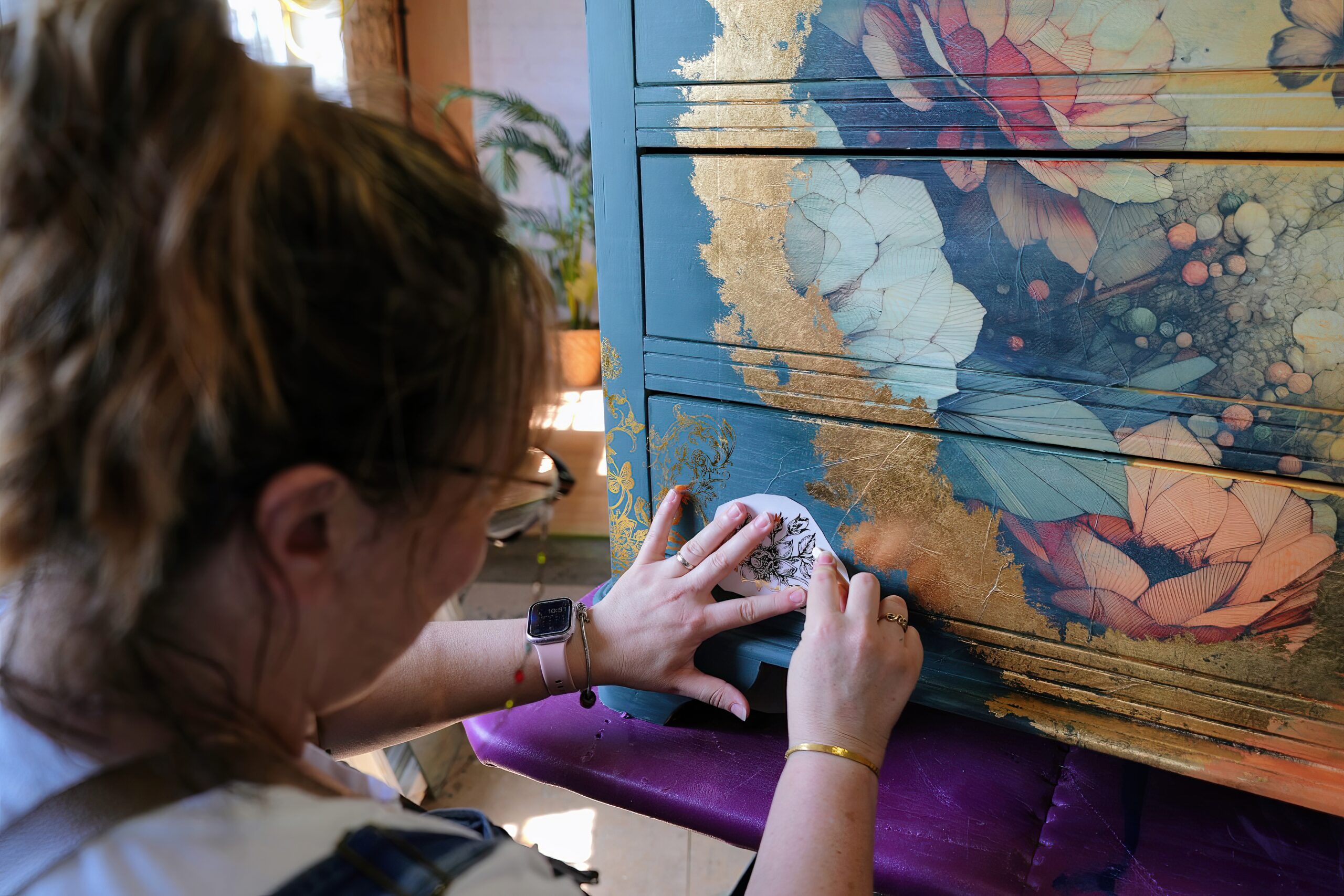 A person with glasses and tied back hair is seen meticulously placing a small floral design on a decorated dresser. The dresser features a vibrant, nature-inspired design with large flowers and gold accents. The person is wearing a smartwatch and working with focus.