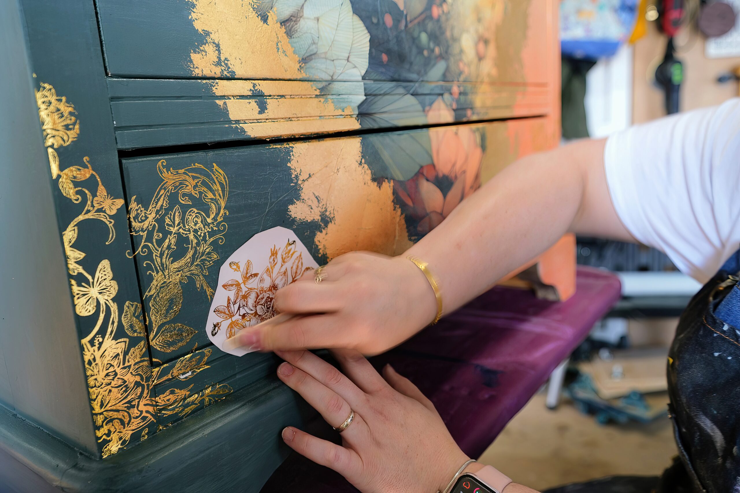 A person applying a floral decal to a green and gold painted piece of furniture. The hands are visible, one wearing a bracelet and the other with a watch. The furniture is decorated in a mix of gold leaf and floral designs.