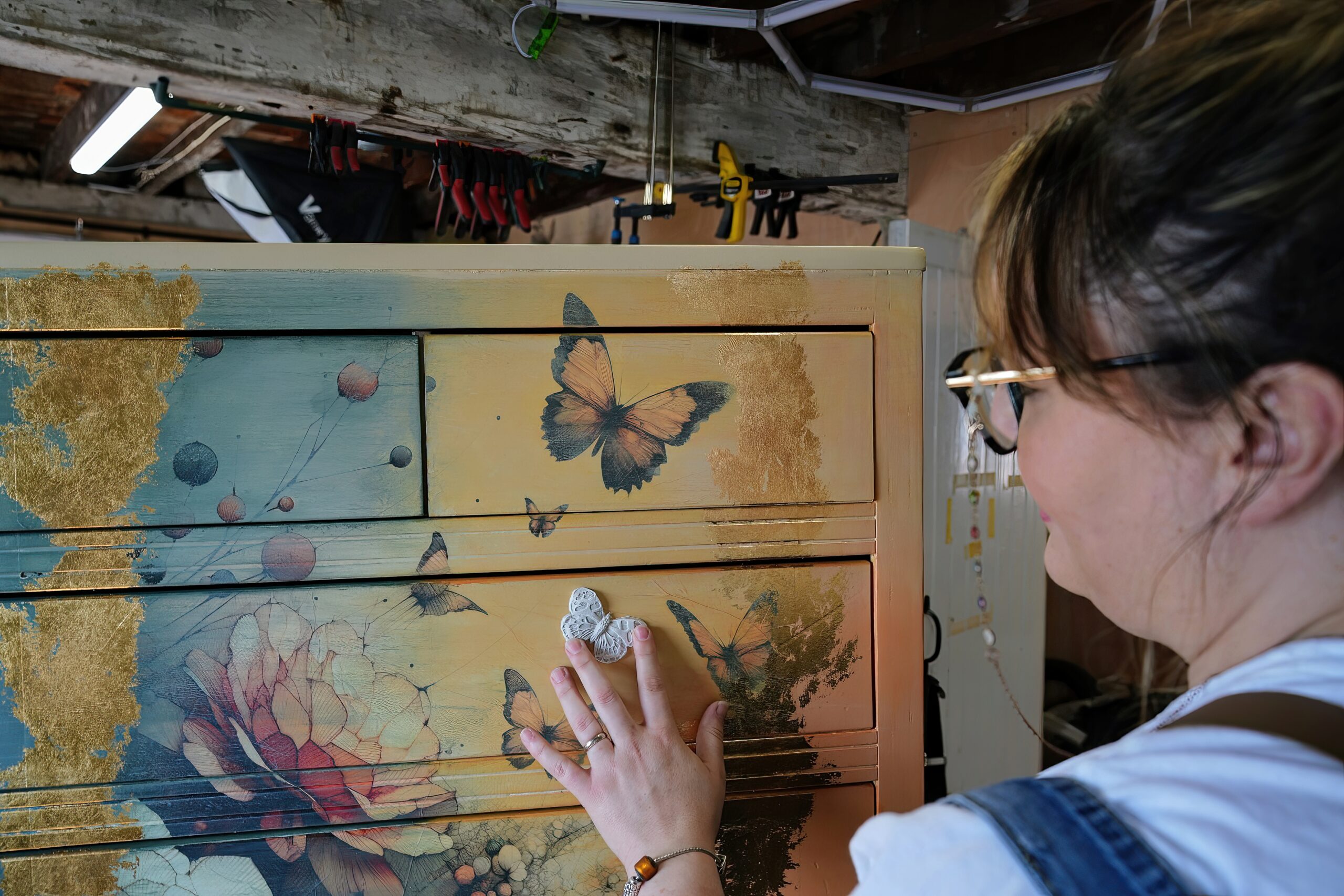 A person with glasses is applying a butterfly decoration to a painted dresser. The dresser has a colorful design featuring flowers and butterflies. The surroundings appear to be a workshop with tools hanging on the wall and visible pipes overhead.