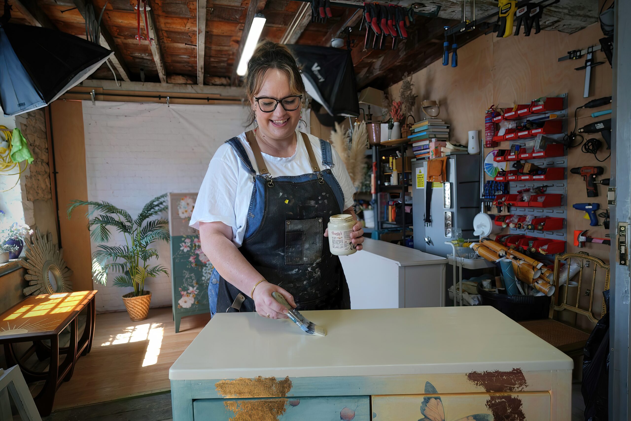 A person wearing glasses and an apron is painting a piece of furniture in a workshop. The workshop is filled with tools, shelves, plants, and various decorative items. The person is smiling and holding a paintbrush, working on the front surface of a dresser.