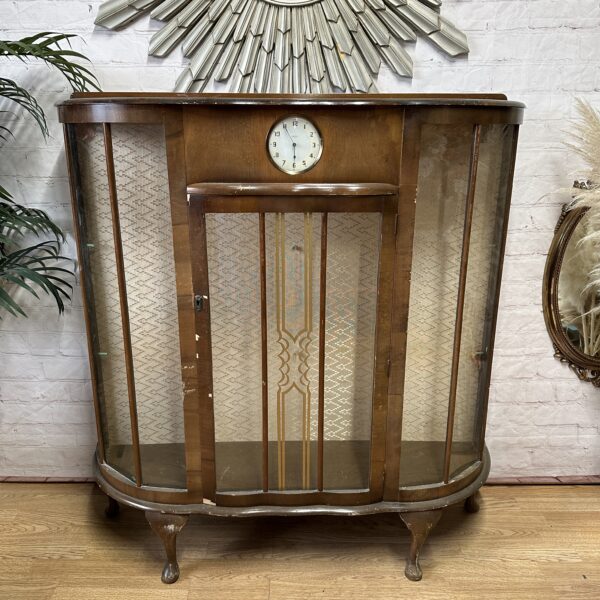 A vintage wooden display cabinet with curved glass sides stands on cabriole legs. It features a clock at the center top and ornate patterned detailing on the front glass panels. Behind it is a brick wall with a decorative round mirror and nearby plants.