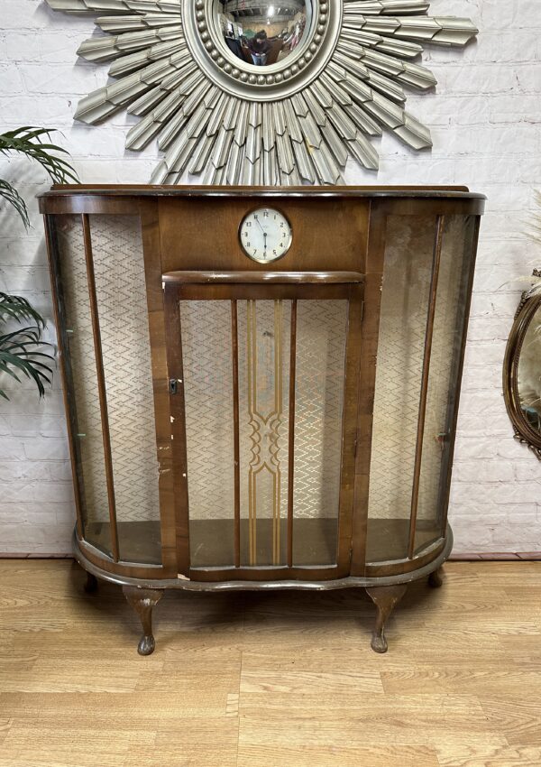 A vintage wooden display cabinet with curved glass sides stands on cabriole legs. It features a clock at the center top and ornate patterned detailing on the front glass panels. Behind it is a brick wall with a decorative round mirror and nearby plants.