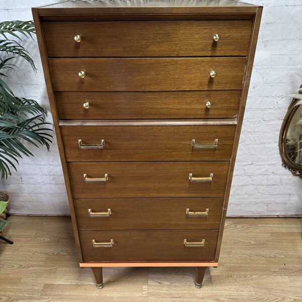 A tall wooden dresser stands against a white brick wall. The dresser has seven drawers, the top three smaller with round knobs and the bottom four larger with rectangular handles. A decorative framed mirror and a green plant are on either side.