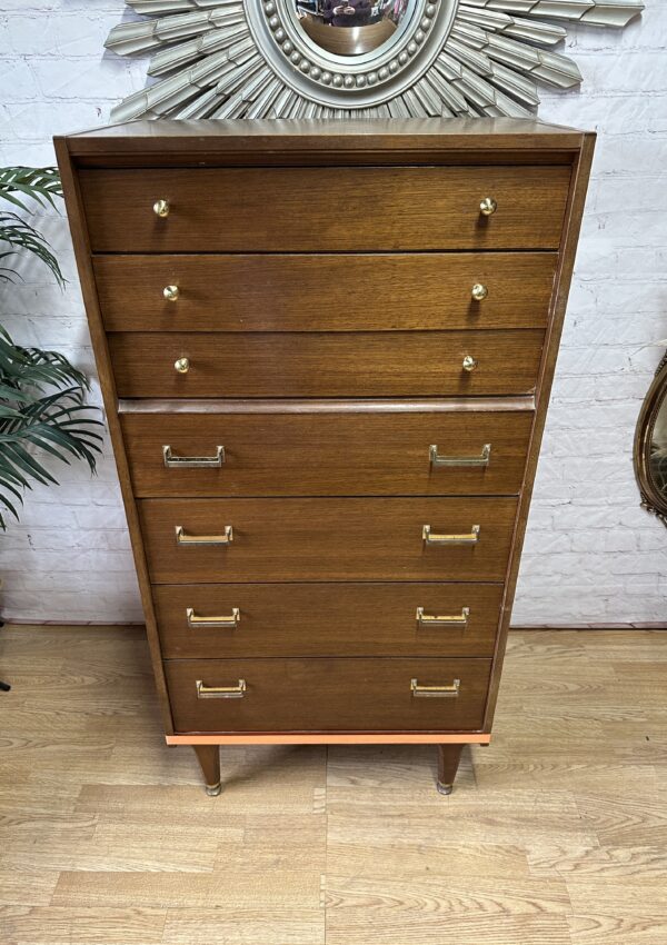 A tall wooden dresser stands against a white brick wall. The dresser has seven drawers, the top three smaller with round knobs and the bottom four larger with rectangular handles. A decorative framed mirror and a green plant are on either side.