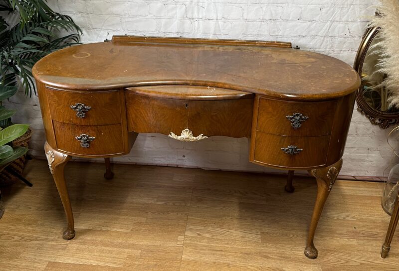 Antique wooden vanity with a curvaceous front, featuring four drawers with intricate metal handles. The vanity has cabriole legs and a light-colored wooden top with visible grain. It stands against a white brick wall, with plants and a decorative mirror nearby.