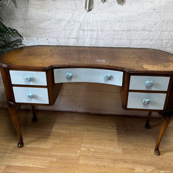 An antique wooden desk with a curved front and six light blue drawers featuring round blue knobs. The desk has cabriole legs and a slightly worn, brown wood finish. It is placed on a wooden floor in front of a white brick wall, surrounded by plants and mirrors.