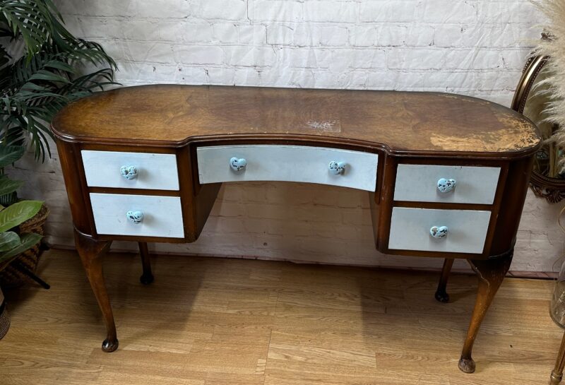 An antique wooden desk with a curved front and six light blue drawers featuring round blue knobs. The desk has cabriole legs and a slightly worn, brown wood finish. It is placed on a wooden floor in front of a white brick wall, surrounded by plants and mirrors.