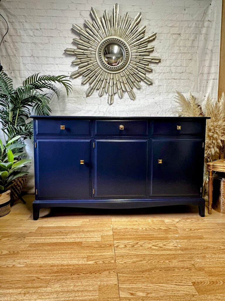 A dark blue sideboard with three drawers and two large cabinet doors sits on a wooden floor. A sunburst mirror hangs above it on a white brick wall. Decorative plants and pampas grass are placed around the sideboard, enhancing the modern, eclectic decor.