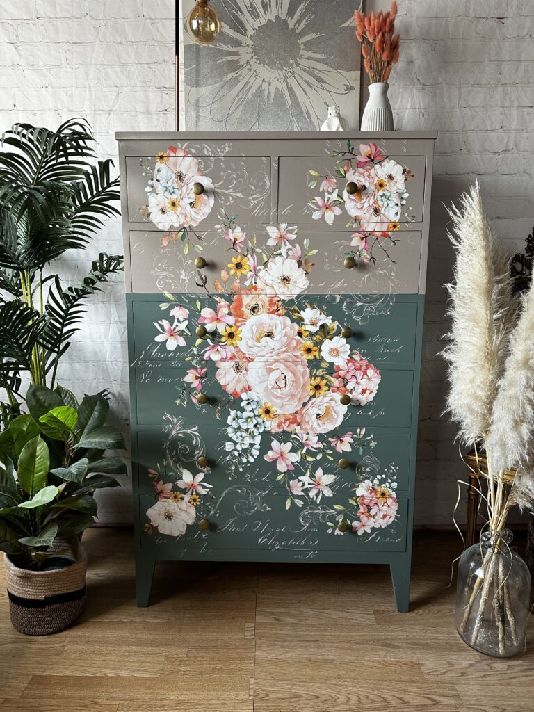 A tall dresser with floral and script designs stands against a white brick wall. The dresser is divided into earthy tones, with pink and white flowers. Surrounding it are various green plants, including potted ferns and pampas grass, adding a natural touch.