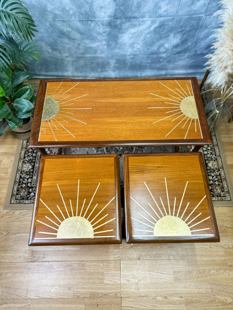 A set of three wooden tables with a polished finish, two smaller ones fitting under a larger one, each adorned with a geometric sunburst design featuring gold accents. The tables are placed on a hardwood floor, with a plant and pampas grass in the background.