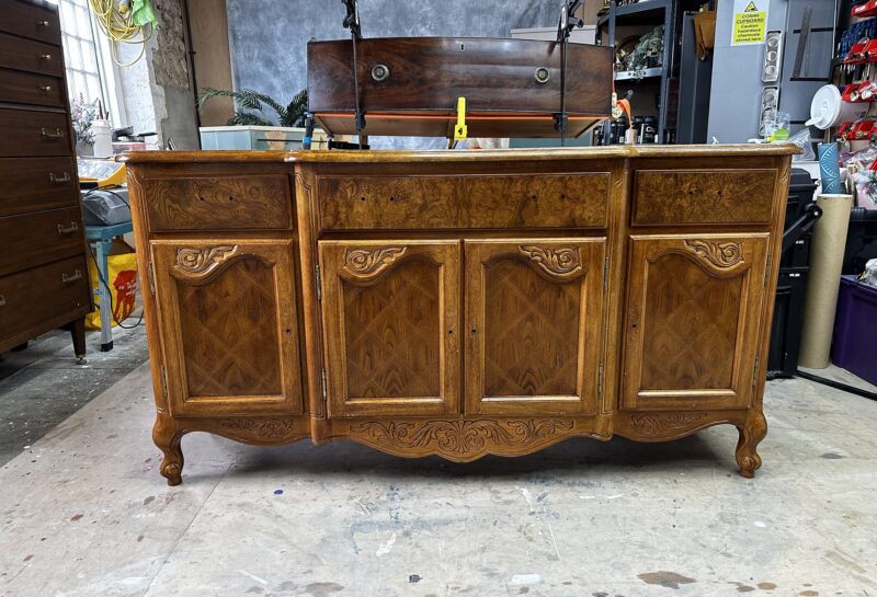 A wooden sideboard with four intricately carved cabinet doors stands in a workshop. The sideboard features ornate detailing and a polished finish. In the background, various tools and workshop equipment are visible on shelves and a workbench.