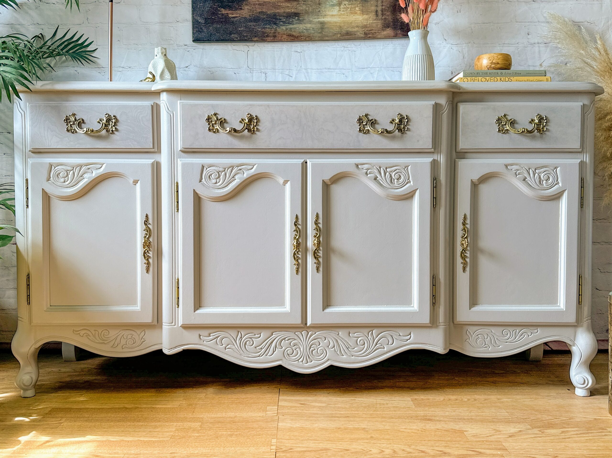 A light beige vintage sideboard with ornate gold handles and intricate carvings stands against a brick wall. The sideboard features three drawers and four cabinet doors. Above it sits a book, a vase with dried flowers, and a decorative item.