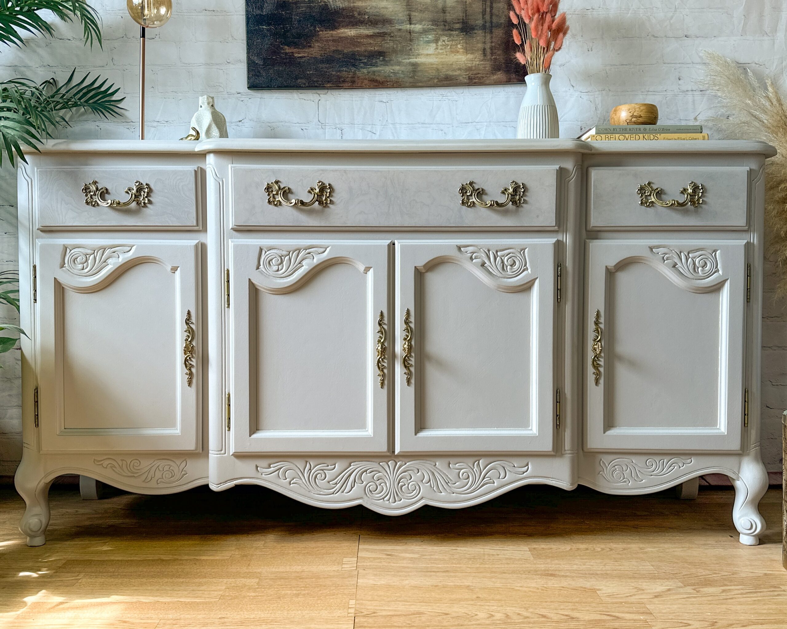 A vintage white sideboard with elaborate gold handles and intricate carvings on its doors and drawers stands on a wooden floor. A book, decor items, and dried flowers are arranged on top, while a piece of painting furniture along with green plants provides a charming backdrop.