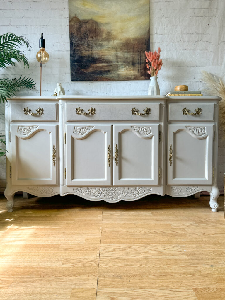 A white vintage-style sideboard with ornate gold handles and accents stands against a white brick wall. It has four doors and two drawers. A painting of a nature scene hangs above it, and decorative items including a lamp, potted plant, and vase sit on top.