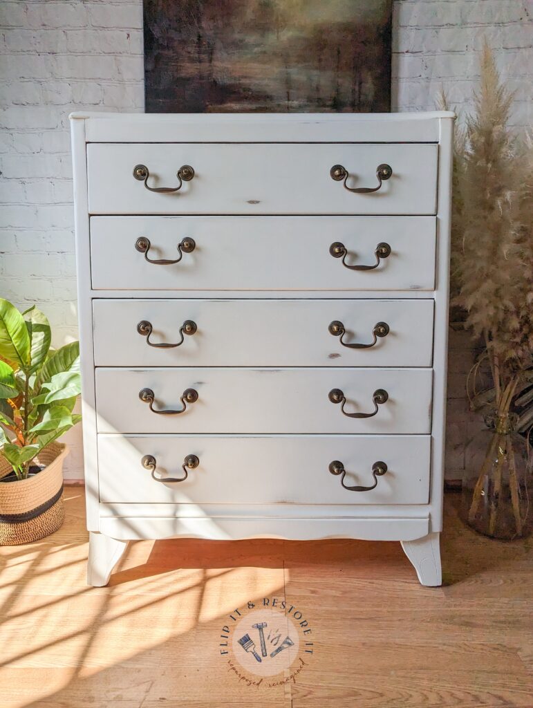 A white wooden dresser with five drawers and vintage-style brass handles stands against a white brick wall. A potted green plant on the left and dried pampas grass on the right accentuate the space. Natural light casts shadows on the dresser and hardwood floor.