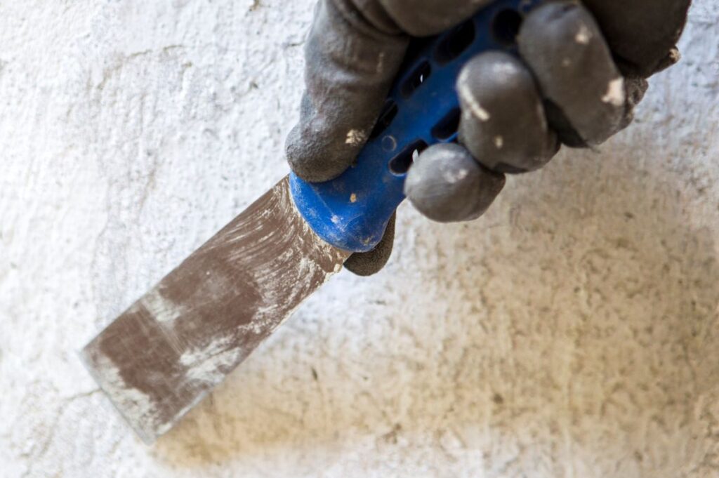 A close-up of a gloved hand using a blue-handled putty knife to scrape a textured white wall. The glove is black with white paint specks, and the putty knife has a metal blade with remnants of dried plaster.