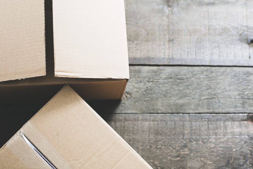 Two cardboard boxes on a wooden floor. The boxes are partially open, revealing their corrugated texture. The wooden floor has a rustic, slightly worn appearance, adding a touch of character to the scene. The image gives a sense of moving or storage.