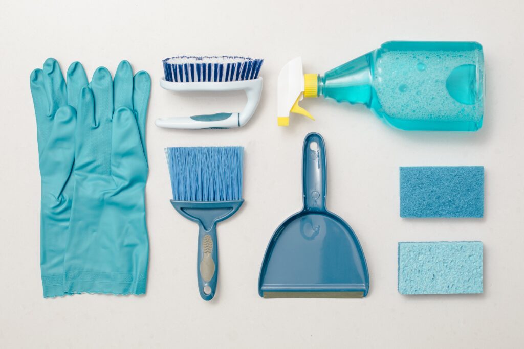 A collection of blue cleaning supplies neatly arranged on a white background. Items include rubber gloves, a scrub brush, a spray bottle with blue liquid, a dustpan and brush, a squeegee, and two sponges.