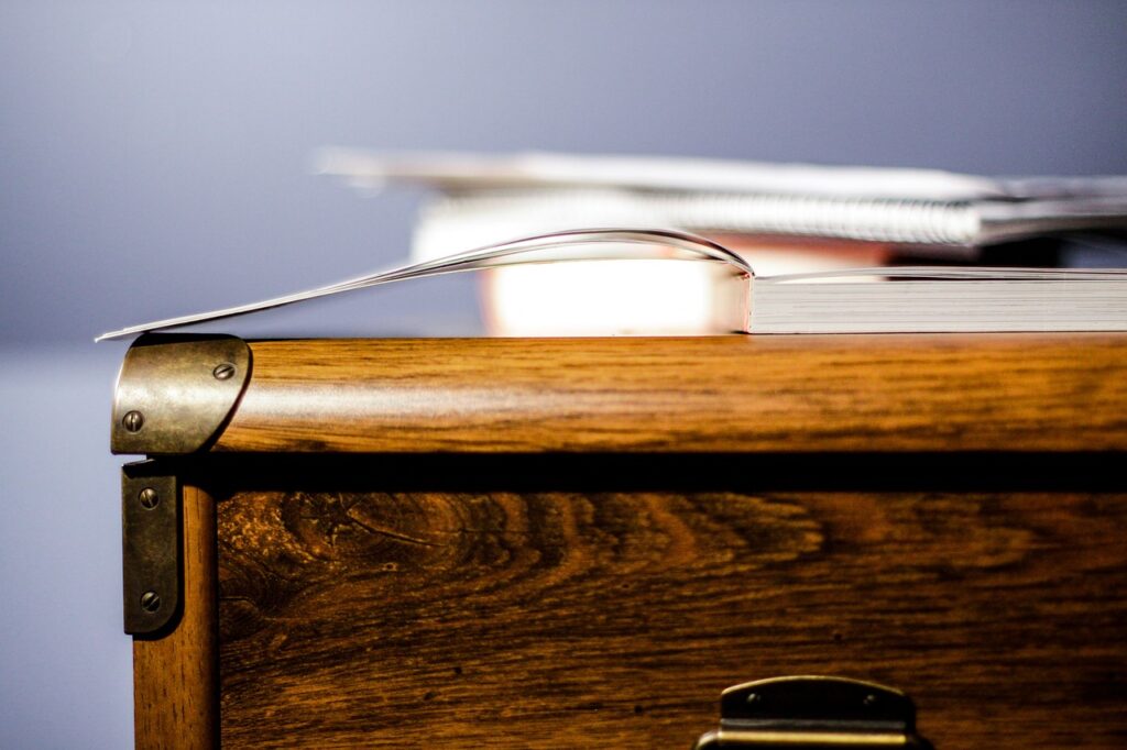 An open book lies on top of a wooden desk, with a spiral notebook partially visible in the background. The desk features ornate metal corner protectors and a latch. The background is blurred, focusing attention on the desk and the book.