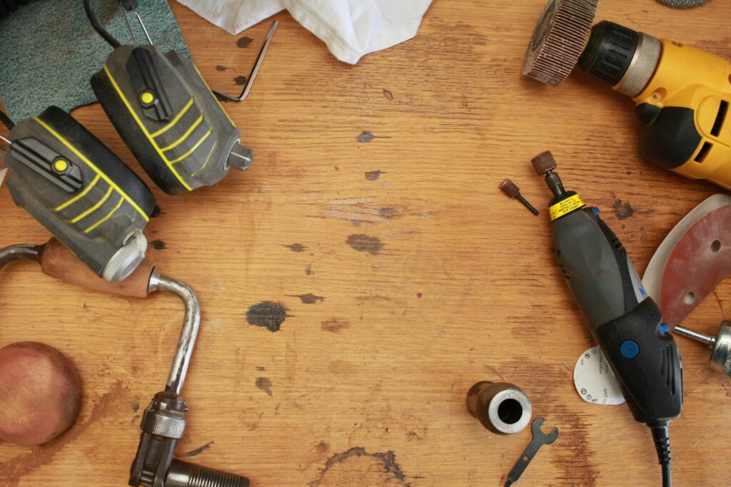 A variety of tools and objects are laid out on a wooden workbench. Visible items include safety goggles, a cordless drill, a rotary tool, a wrench, and several other small tools and hardware. The center of the workbench is mostly clear, showing a worn surface.