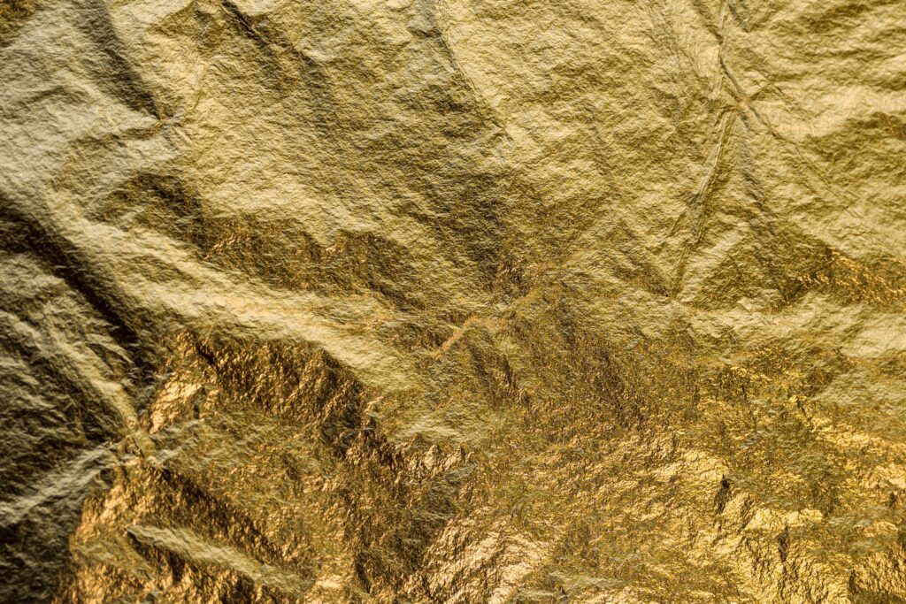 A close-up image of crinkled gold foil. The surface has a textured, uneven appearance, with varying shades of gold that catch the light, creating a shimmering effect. The foil is characterized by its metallic shine and intricate folds.