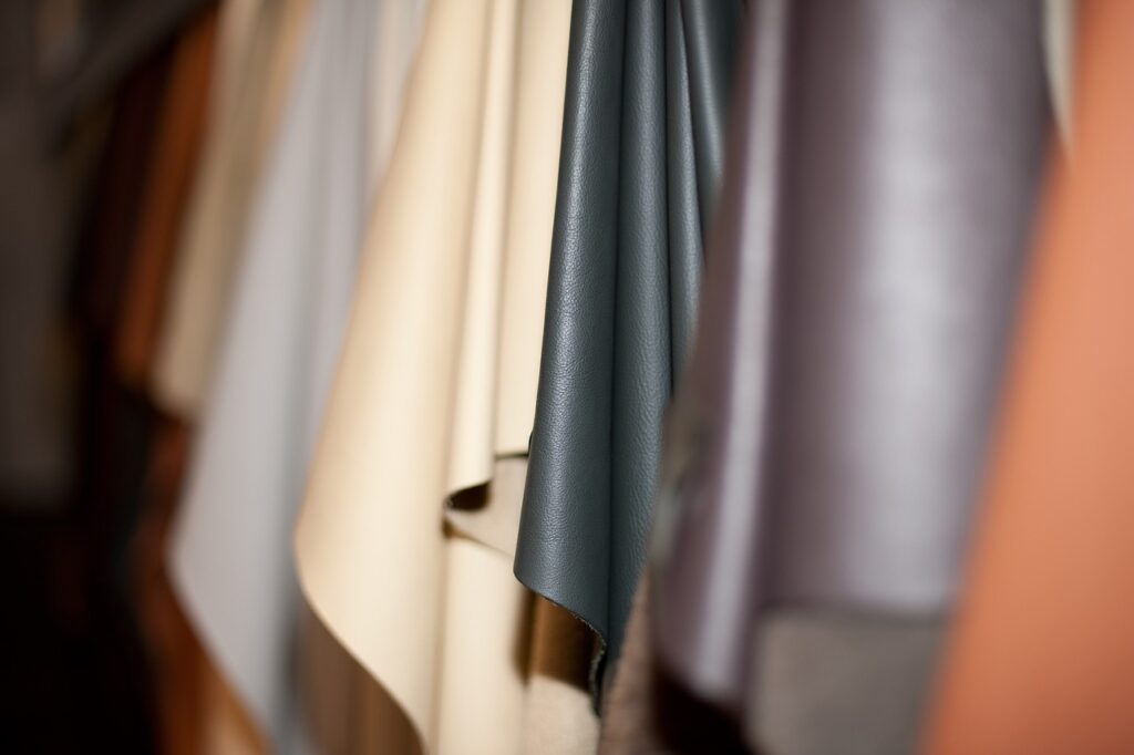 Close-up image of various shades of leather fabric hanging in a row. The colors range from light beige to dark blue and brown, showcasing different textures and finishes. The focus is primarily on the blue leather piece, with others slightly blurred.