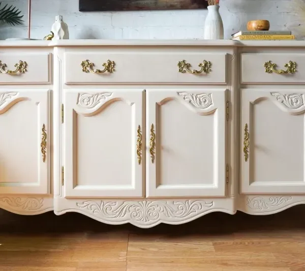 A vintage-style, cream-colored wooden dresser with ornate carvings and gold handles. It features three drawers on top and two large cabinets below. The dresser stands on curved legs and is placed against a white brick wall with decorative items on top.