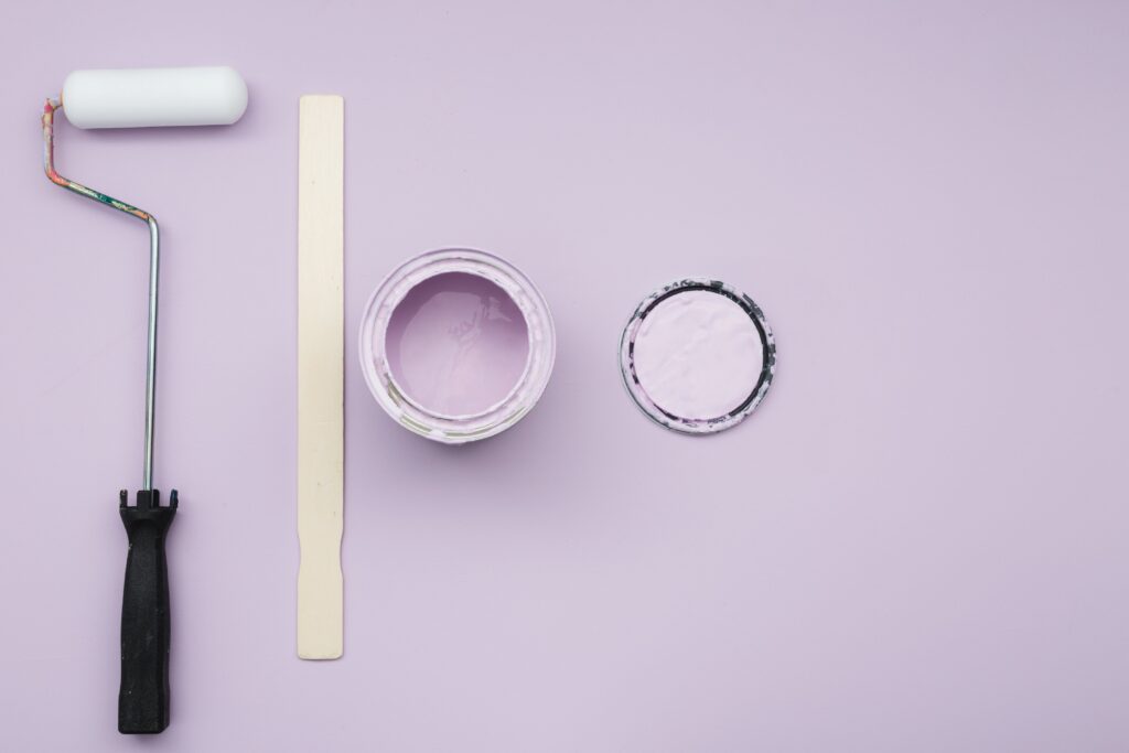 A paint roller, a wooden stirring stick, and an open can of light purple paint with its lid placed beside it are arranged on a light purple background. The roller is on the left, the stirring stick is in the center, and the paint can and lid are on the right.