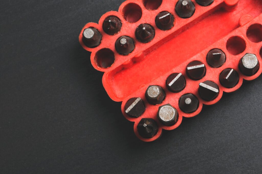 A close-up image of a red plastic case containing several different types of screwdriver bits. The case is open, revealing the metallic bits, each designed for various screw head types, including flathead and Phillips. The background is dark and contrasts with the red case.