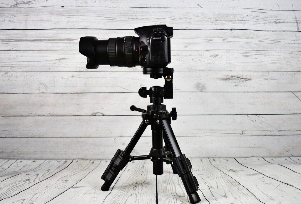 A professional DSLR camera with an attached lens is mounted on a compact, adjustable tripod. The background and floor are made of white wooden planks, providing a rustic and clean setting. The camera and tripod are black and appear ready for use.